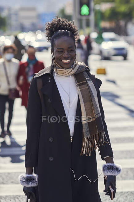 Jovem feliz na moda afro-americana com mochila e óculos de sol passeando na estrada urbana enquanto olha para a câmera à luz do sol — Fotografia de Stock
