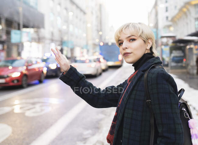 Positive junge Frau in warmem Mantel hebt den Arm, um an einem Wintertag in Madrid ein Taxi auf einer belebten Straße zu begrüßen — Stockfoto