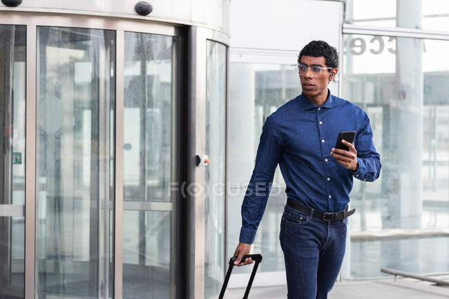 Дорослий етнічний чоловік у розумному повсякденному одязі з мобільним телефоном і багажем, який дивиться у далечінь під час подорожі — стокове фото