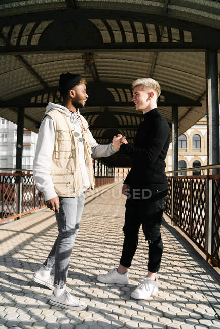 Side view of stylish young diverse male friends in trendy outfits standing on paved street in city and shaking hands — Stock Photo