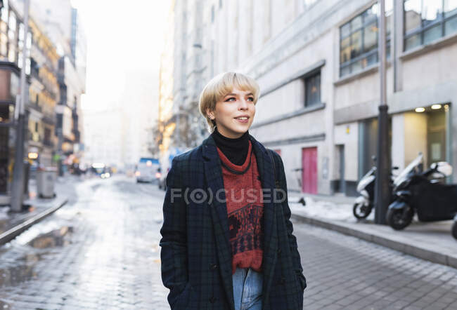 Belle femme positive en tenue tendance et manteau chaud debout avec les mains dans les poches sur la rue enneigée de la ville et regardant loin par temps clair d'hiver à Madrid, Espagne — Photo de stock