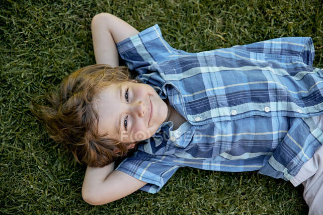 Vue du dessus du petit enfant heureux en chemise à carreaux souriant et regardant la caméra tout en étant couché sur la pelouse avec les mains derrière la tête — Photo de stock