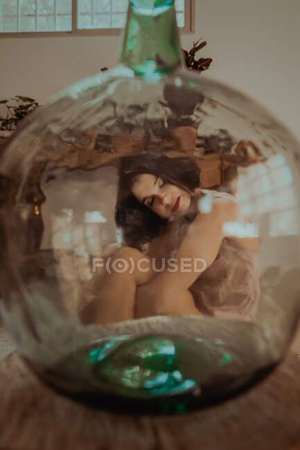 A través de una botella de vidrio de delicada hembra sentada en el suelo y abrazando las rodillas en la habitación en casa - foto de stock