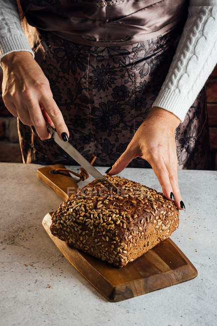 Cultivo de panadero femenino anónimo con cuchillo de corte de pan fresco con semillas de girasol en la mesa en panadería - foto de stock