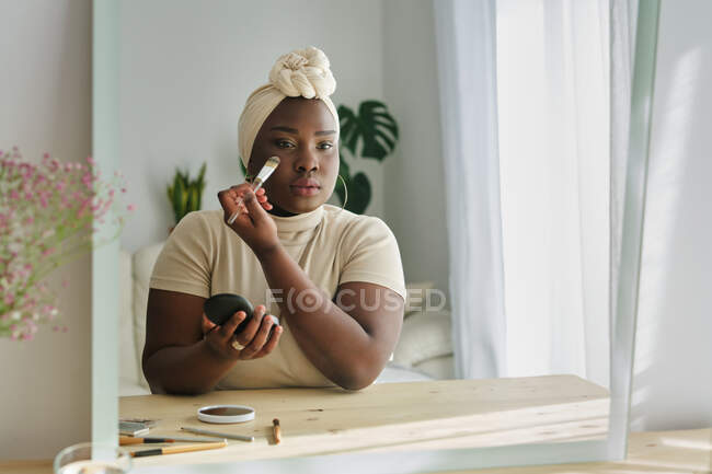 Stilvolle junge, klobige Afrikanerin in traditionellem Turban, die im hellen, modernen Raum neben Spiegel steht und Foundation auf das Gesicht aufträgt — Stockfoto