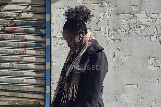 Vista lateral de una joven mujer étnica atenta en bufanda con moño de pelo afro escuchando canciones en auriculares en la ciudad mientras mira hacia abajo - foto de stock