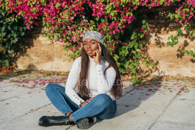 Entschlossene Afroamerikanerin mit langen Haaren und lässigem Outfit sitzt an sonnigen Tagen im Park und blickt in die Kamera — Stockfoto