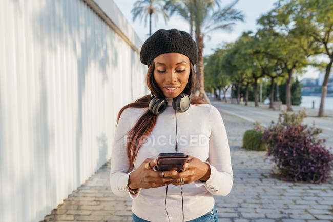 Deliziosa femmina nera con cuffie sul collo appoggiate alla costruzione e alla navigazione del telefono cellulare sulla strada della città — Foto stock