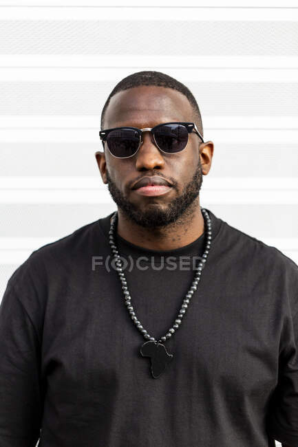 Unemotional calm African American man in black t shirt with Africa continent necklace and sunglasses standing looking at camera on light background — Stock Photo