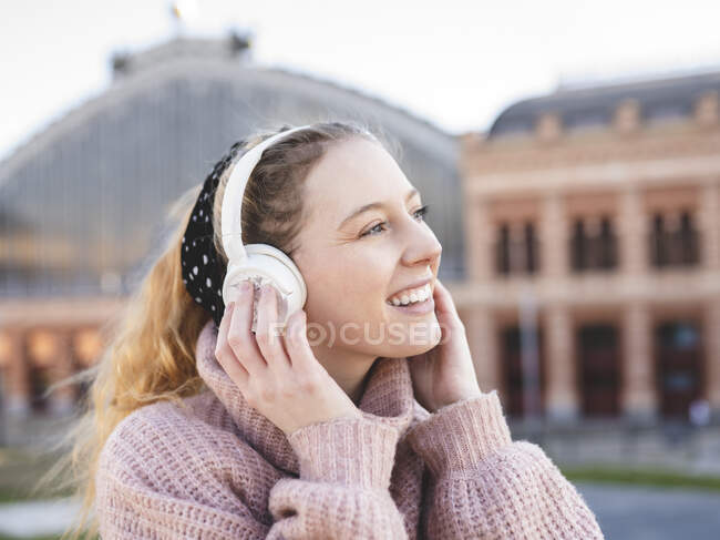 Side view of cheerful teenage female with long wavy hair in warm sweater smiling while enjoying music in headphones standing on city square — Stock Photo