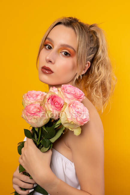 Молода неемоційна красива жінка в білій сукні з голими плечима, що тримає ніжні рожеві троянди, стоячи, дивлячись на камеру на жовтому тлі в фотостудії — стокове фото
