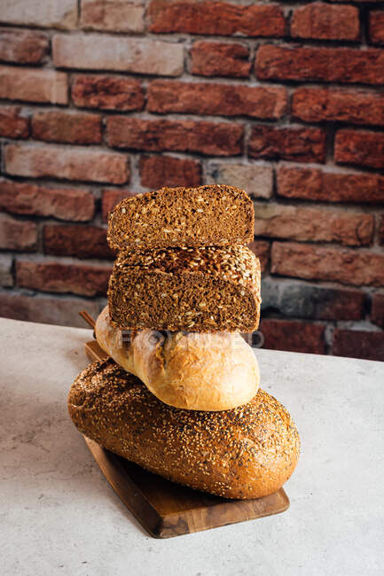 Pilha de pão branco e de centeio com cereais e crosta apetitosa na tábua de corte contra a parede de tijolos na padaria — Fotografia de Stock