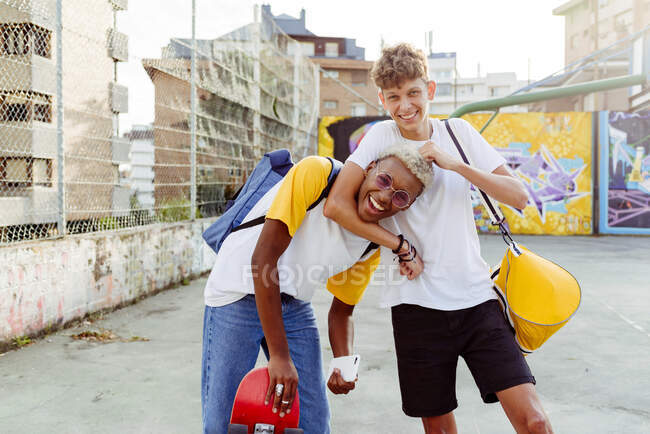 Два подростка со скейтбордом и рюкзаком обнимаются и смеются на улице. — стоковое фото