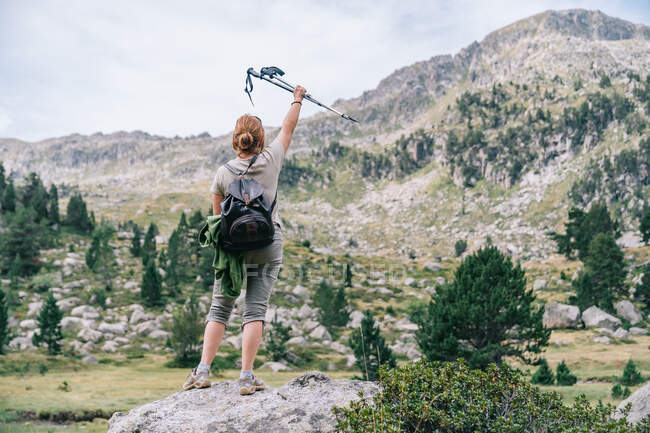 Back view anonymous female hiker in casual clothes with backpack raising hand with Nordic walk poles while standing on stony hilltop in mountainous Ruda Valley in Catalan Pyrenees — Stock Photo