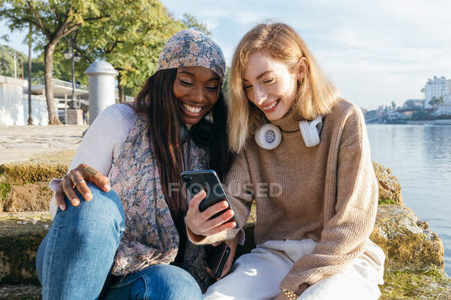 Посміхаючись, багаторасові подружки сидять на набережній у сонячний день і спілкуються на смартфоні. — стокове фото