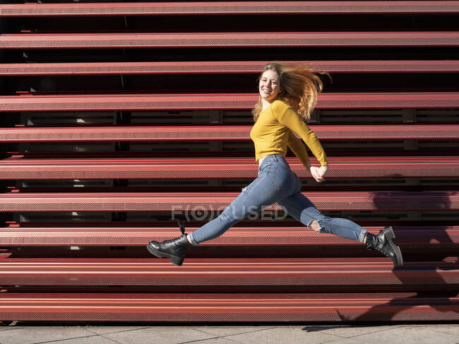 Full body of cheerful young female millennial in casual outfit jumping and smiling near metal fence on street — Stock Photo