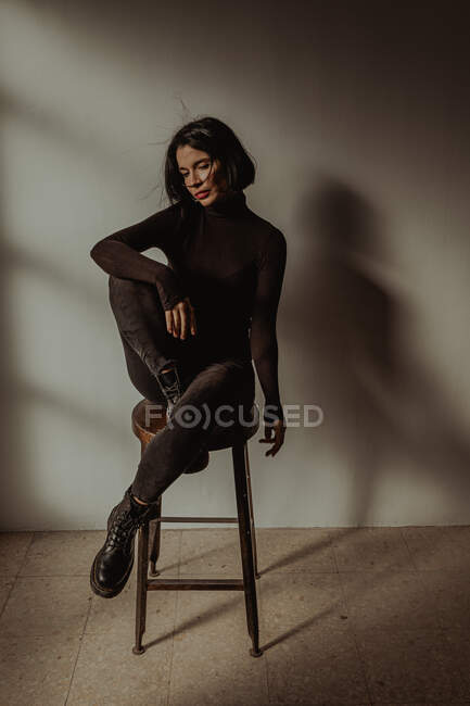 Slim female in black outfit sitting on wooden stool with raised legs in room against white wall — Stock Photo
