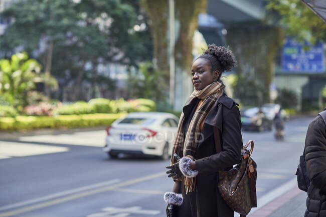 Joven mujer étnica de moda en abrigo y bufanda con moño de pelo afro mirando hacia adelante en el pavimento urbano - foto de stock