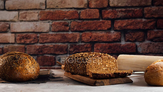 White and rye bread with cereals and appetizing crust on cutting board against brick wall in bakehouse — Stock Photo