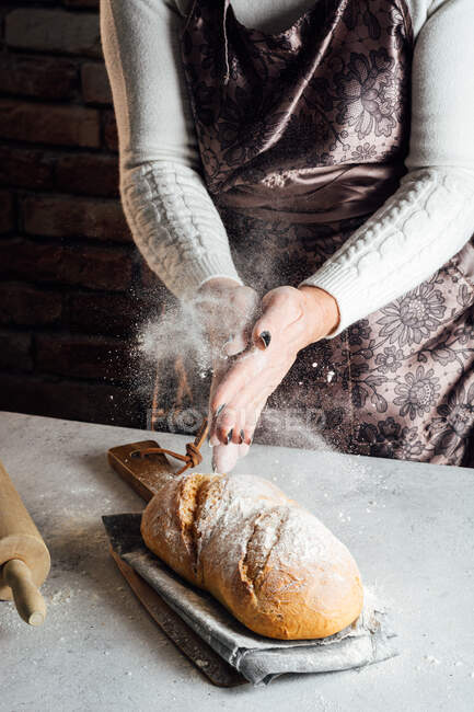 Crop anonymous female artisan in apron sprinkling delicious loaf of white bread with flour at table in bakehouse — Stock Photo