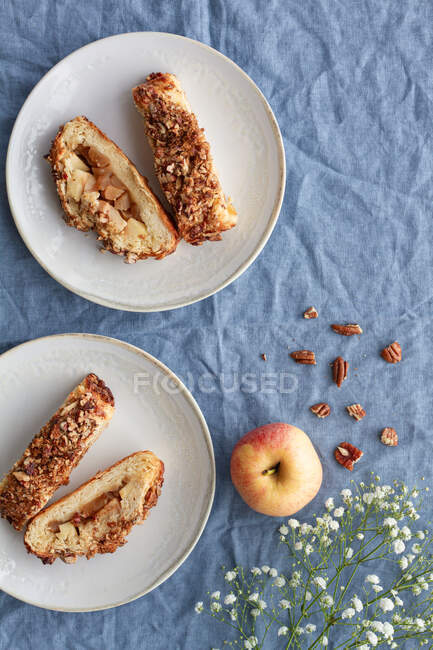 Overhead view of yummy apple pastry on ceramic saucers near fresh fruit and crunchy pecan nuts with flower on creased textile — Stock Photo