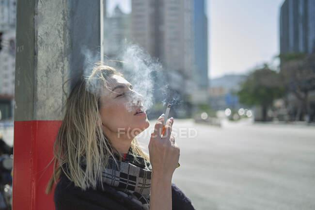 Vista lateral de la joven hembra en bufanda fumar cigarrillo cerca de poste en la carretera urbana en la parte posterior iluminada - foto de stock