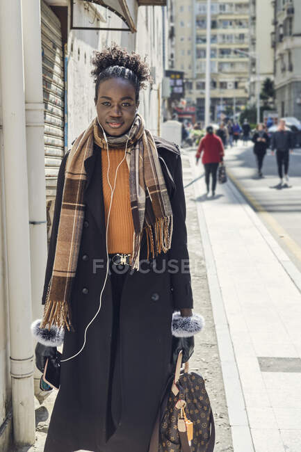 Young trendy African American female with rucksack and sunglasses strolling on urban roadway while looking at camera in sunlight — Stock Photo