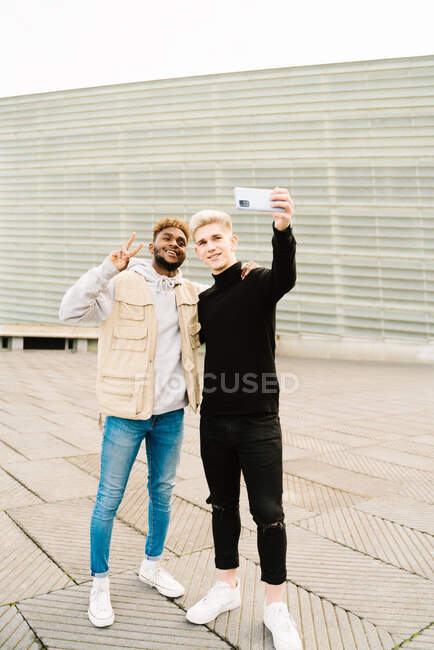 Happy young African American guy in trendy clothes smiling while making a self portrait on street with hand on shoulder of male friend — Stock Photo