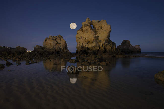 Живописный вид на пляж Прибытия с скальными формациями в чистом рябком море под облачным небом с луной ночью в Алгарве, Португалия — стоковое фото