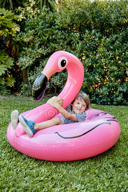 Полное тело веселого маленького мальчика в повседневной одежде, сидящего на надувном розовом фламинго, развлекаясь на травянистой лужайке в парке — стоковое фото
