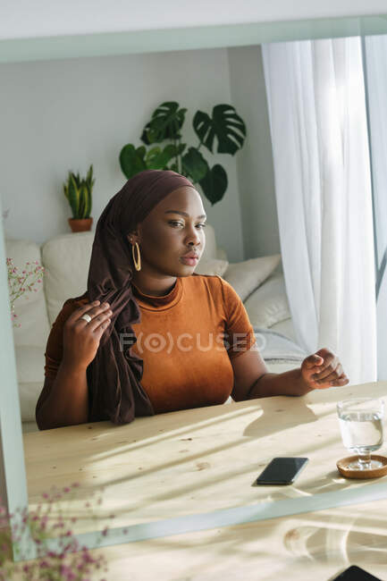 Confiant jeune femme africaine élégante dans le hijab traditionnel appuyé sur la table cosmétique et regardant dans le miroir à la lumière du soleil à la maison — Photo de stock