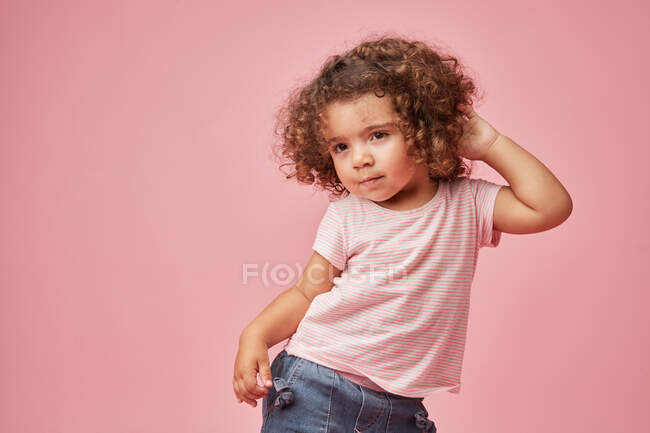 Adorable enfant en bas âge avec les cheveux bouclés regardant loin debout posant avec les mains sur la tête sur fond rose — Photo de stock