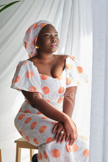 Retrato de jovem senhora africana em vestido de verão elegante com turbante tradicional enquanto sentado perto da janela na sala de luz — Fotografia de Stock
