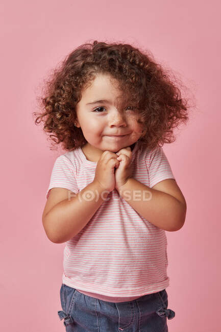 Mignon heureux tout-petit fille avec des cheveux bouclés dans des vêtements décontractés en regardant la caméra sur fond rose — Photo de stock