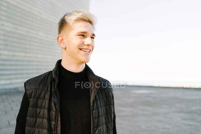 Crop of happy young male in trendy outfit standing on paved street and looking away on sunny day — Stock Photo