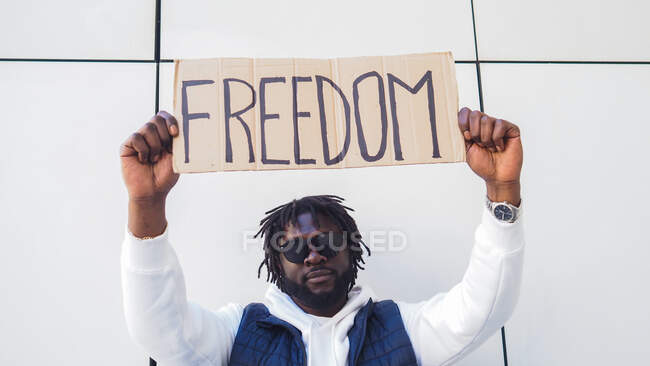 Hombre afroamericano guerrero de justicia social en gafas de sol levantando pancarta de cartón con inscripción Freedom durante la manifestación - foto de stock