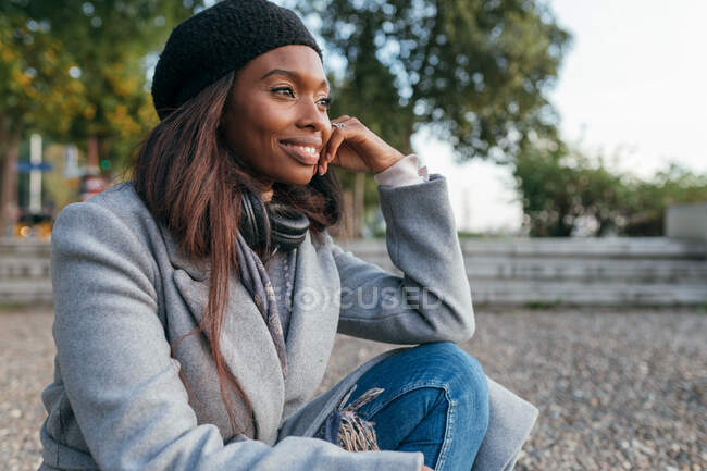 Donna afroamericana spensierata in capispalla seduta sulla strada e appoggiata a portata di mano mentre distoglie lo sguardo dalla contemplazione — Foto stock