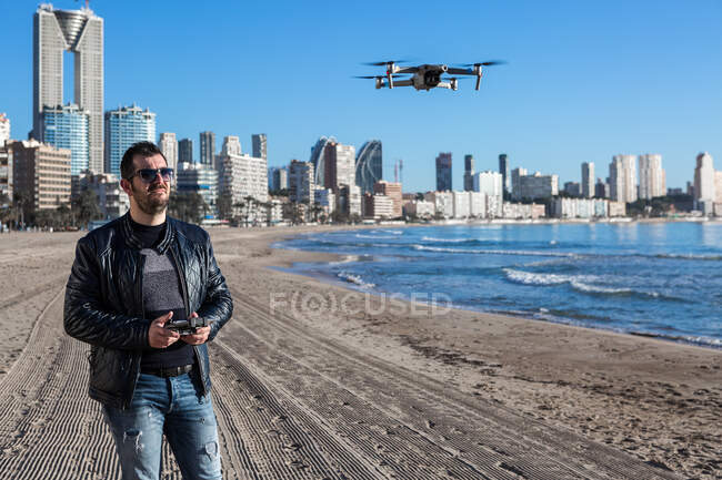 Varón serio en gafas de sol con mando a distancia parado en el avión no tripulado de vuelo de playa en la costa cerca de la ciudad - foto de stock