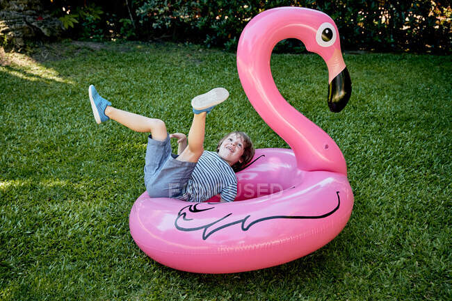 Ganzkörper fröhlicher kleiner Junge in lässiger Kleidung auf aufblasbarem rosa Flamingo liegend, während er sich auf der Rasenfläche im Park vergnügt — Stockfoto