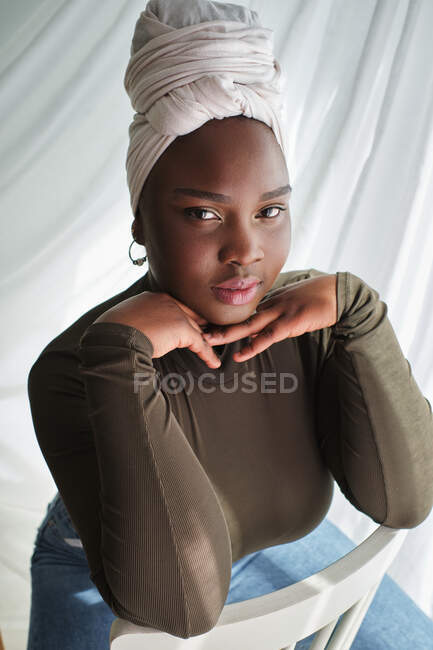 From above of alluring young black lady in casual clothes and turban sitting on chair with hands at chin and looking at camera — Stock Photo