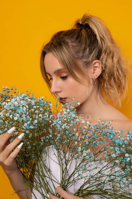 Atractiva joven hembra en vestido blanco con hombros desnudos de pie con flores de gypsophila tiernas azules sobre fondo amarillo en el estudio - foto de stock