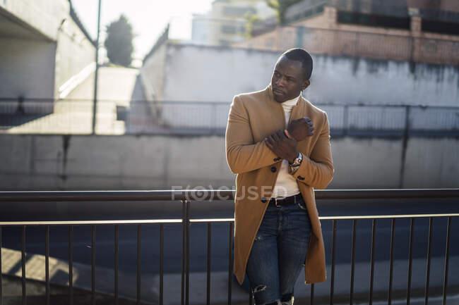 Концентрированный молодой этнический мужчина в модном наряде, опирающийся на забор на улице в солнечный день — стоковое фото