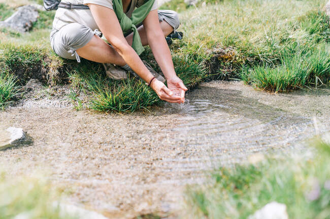 Corpo inteiro cortado mochileiro feminino irreconhecível em uso casual lavar as mãos em riacho enquanto sentado no prado gramado em terreno pedregoso — Fotografia de Stock