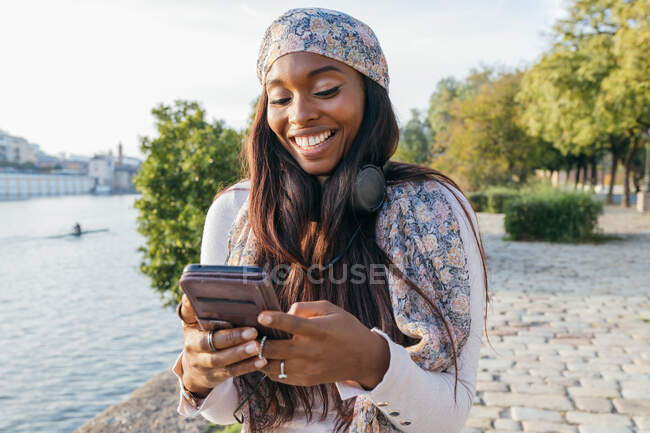 Positive Afroamerikanerin, die an sonnigen Tagen auf der Promenade in der Stadt steht und in den sozialen Medien per Smartphone kommuniziert — Stockfoto