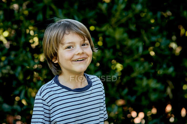 Joyeux petit enfant en chemise à carreaux souriant et regardant la caméra tout en se tenant debout sur la pelouse — Photo de stock