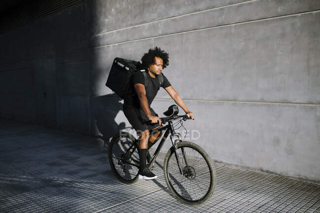 From above of ethnic male with delivery box riding bicycle on walkway against wall with shadow in sunlight — Stock Photo