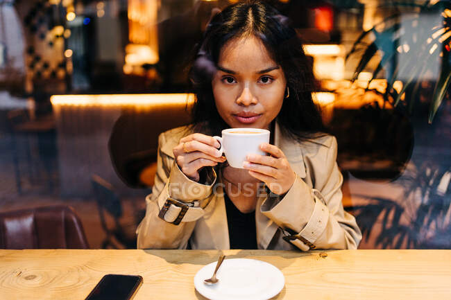 Длинноволосая брюнетка азиатка пьет кофе в кофейне, пока смотрит на сотовый телефон — стоковое фото