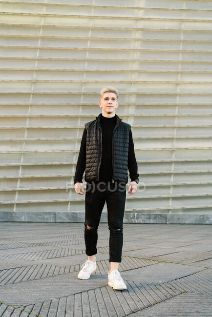 Полное тело самоуверенного молодого мужчины в модном наряде и кроссовках, стоящего на асфальтированной улице и смотрящего в солнечный день — стоковое фото