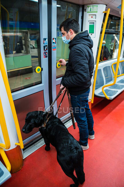 Side view full length of male passenger in mask with eyesight disability standing near doors of subway wagon with guide dog — Stock Photo