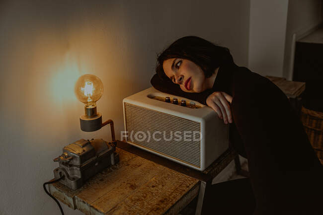 Alto ángulo de soñadora hembra apoyada en la radio en la habitación retro y mirando la bombilla iluminada - foto de stock